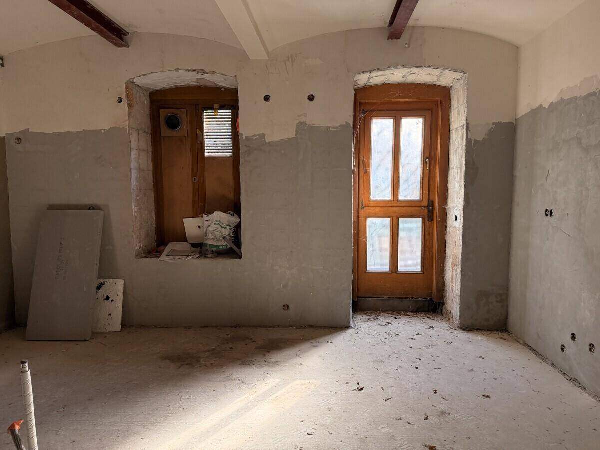 Renovierungsraum mit unverputzten Wänden, einem Holztürrahmen und einem Fenster, das auf den Außenbereich blickt und Licht spendet.
