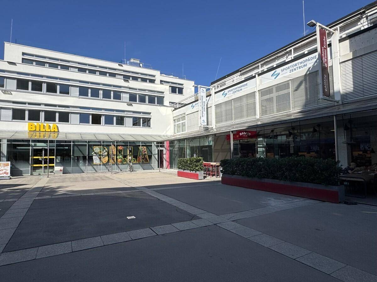 Außenansicht eines modernen Geschäftskomplexes mit verschiedenen Läden und einem sonnigen Platz.