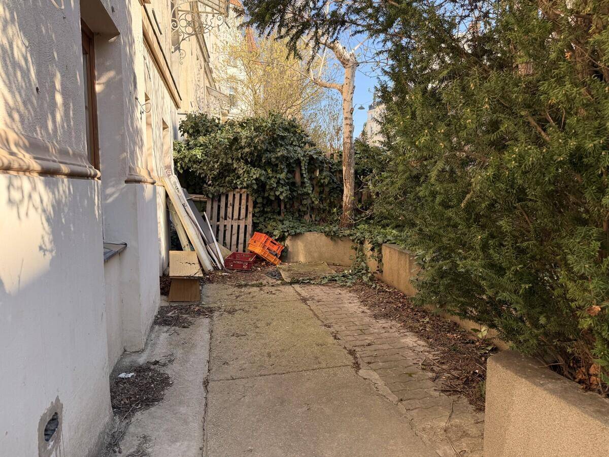 Vernachlässigter Außenbereich mit Betonweg, umgeben von Sträuchern und einer Mauer, neben einem Gebäude mit älterer Architektur.
