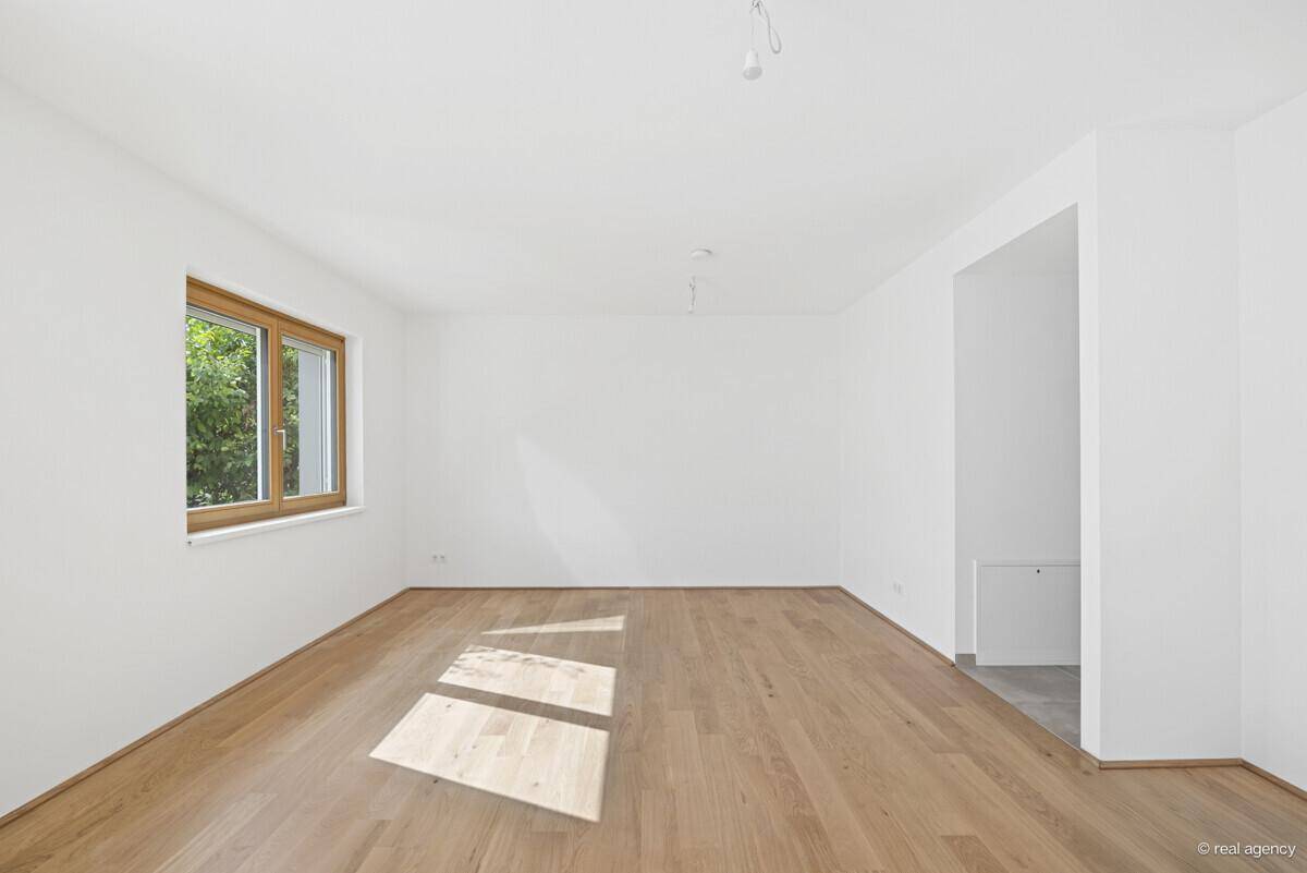Leerer, heller Raum mit Holzboden und großem Fenster, das viel Tageslicht hereinlässt.