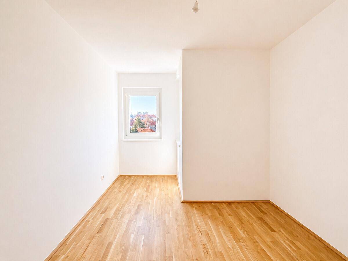 Heller, leerer Raum mit Holzboden und einem Fenster, das viel Tageslicht hereinlässt.