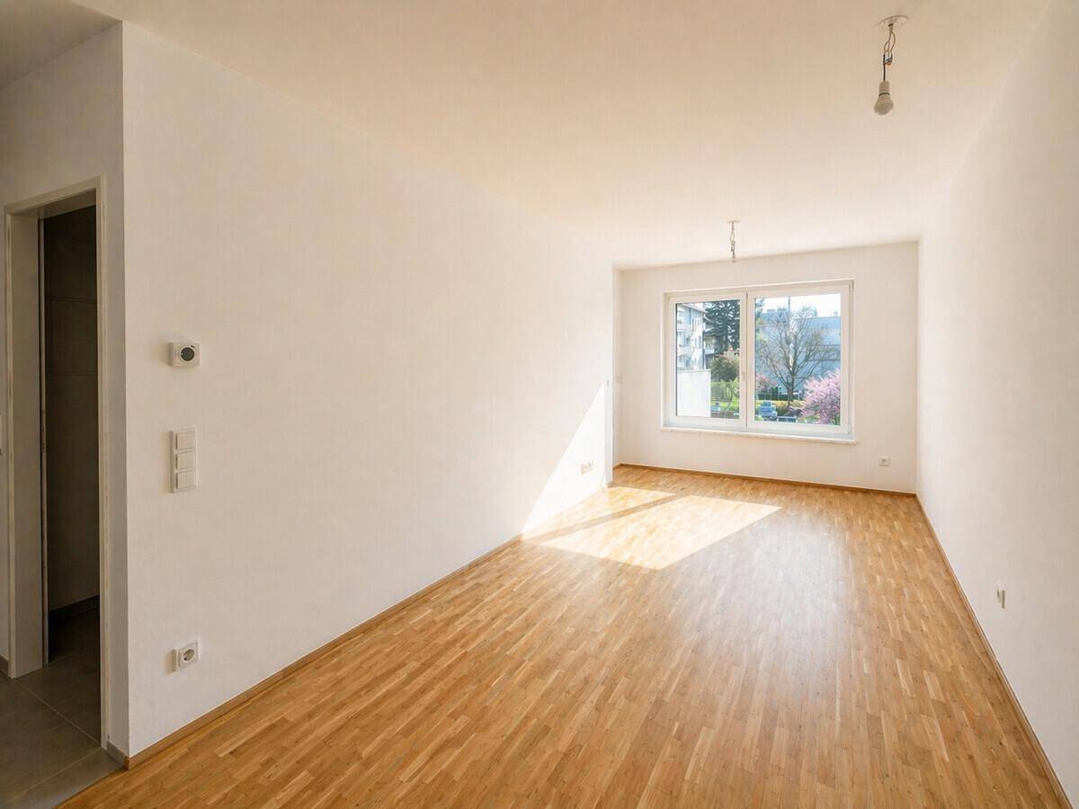 Helles, leeres Zimmer mit Holzparkettboden und einem Fenster, das viel Tageslicht hereinlässt.