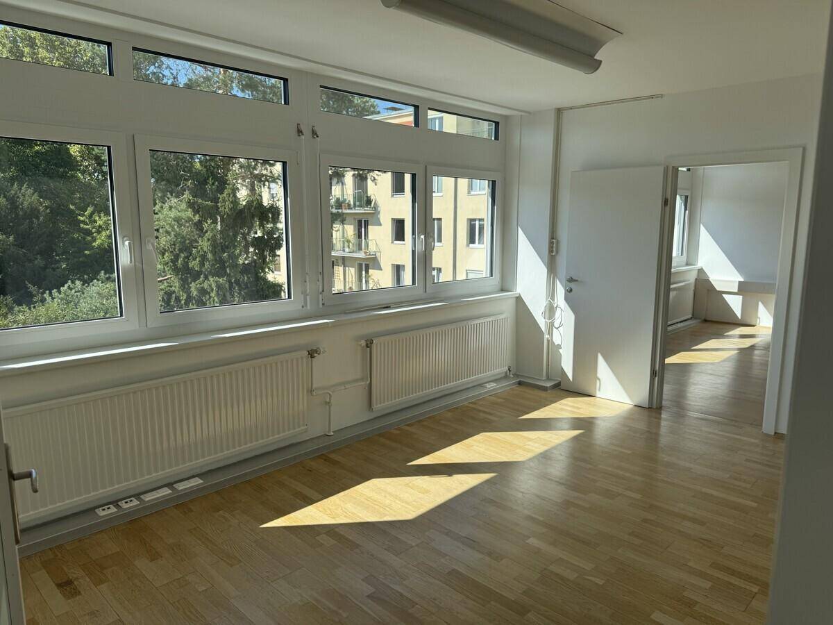 Geräumiges, helles Büro mit Holzboden und großen Fenstern, die einen Ausblick auf Bäume und Gebäude ermöglichen.