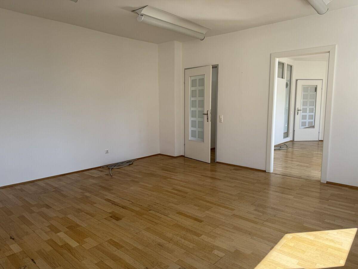 Heller, leerer Büroraum mit Holzboden und weißen Wänden, der Zugang zu weiteren Räumen bietet.