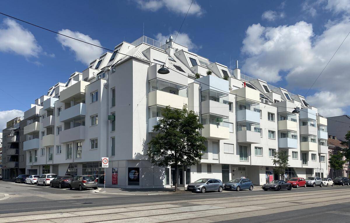 Modernes Mehrfamilienhaus mit weißer Fassade, vielen Balkonen und großen Fenstern an einer Straßenecke.