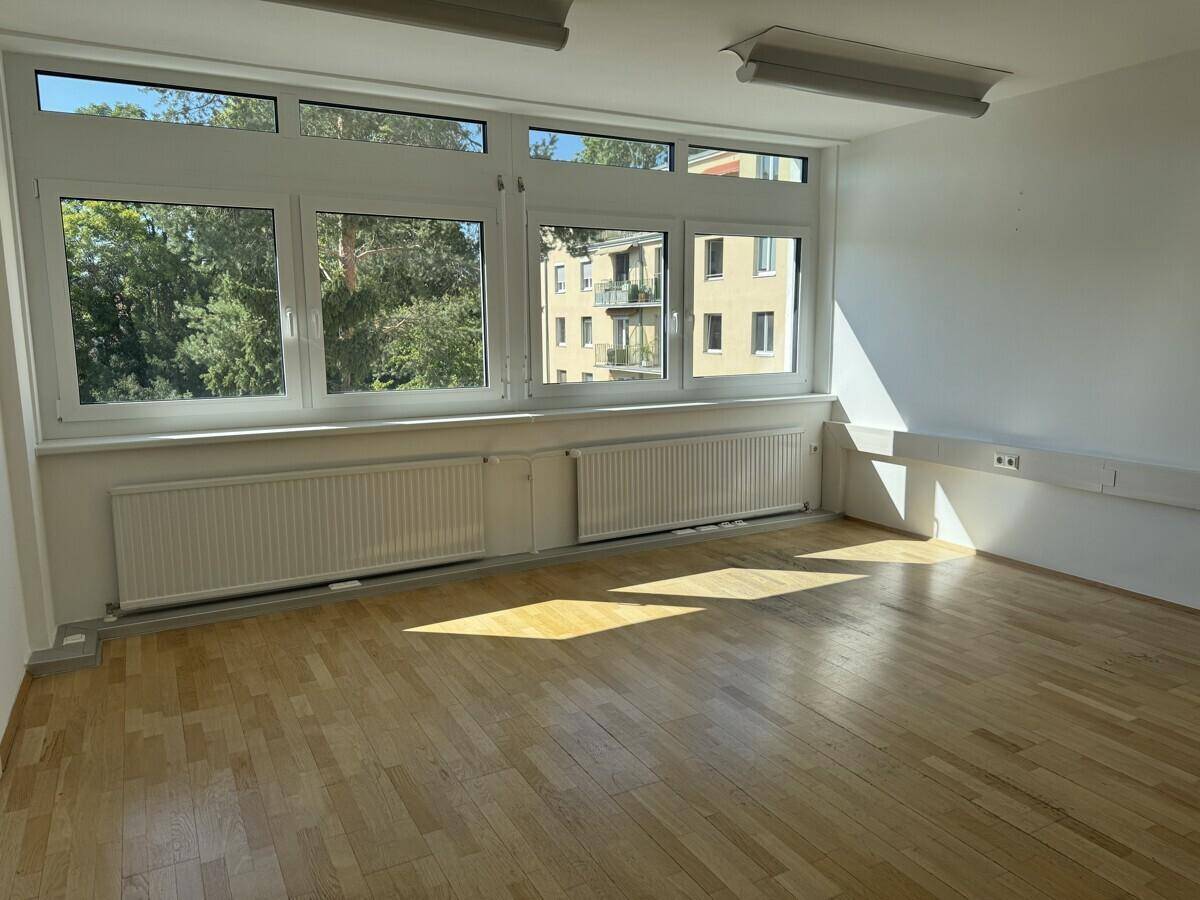 Leerer Büroraum mit Holzboden und großen Fenstern, die viel Sonnenlicht und einen Blick ins Grüne bieten.