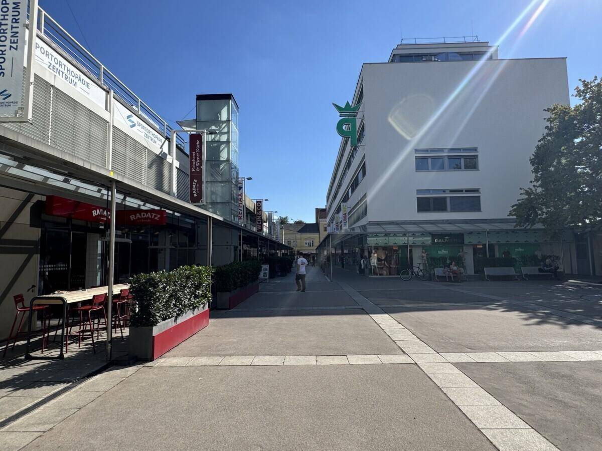 Fußgängerzone mit Geschäften und Restaurants, umgeben von modernen Gebäuden unter blauem Himmel.