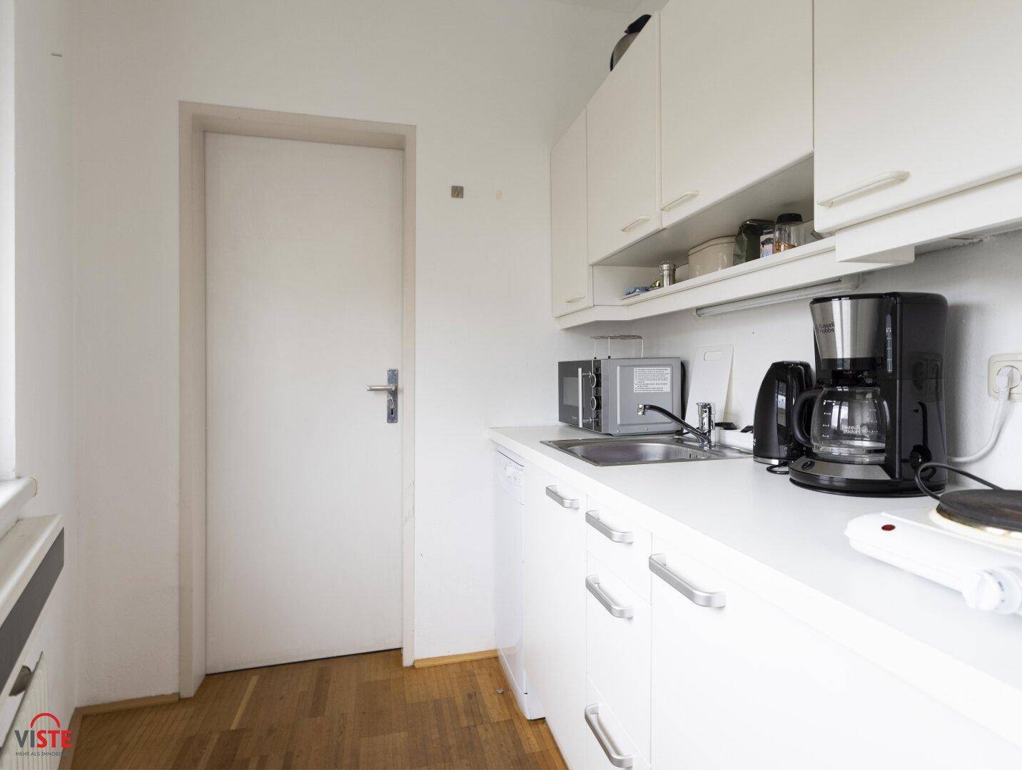 Kleine Küche mit weißen Schränken, Spüle und verschiedenen Elektrogeräten, ideal als Teeküche.