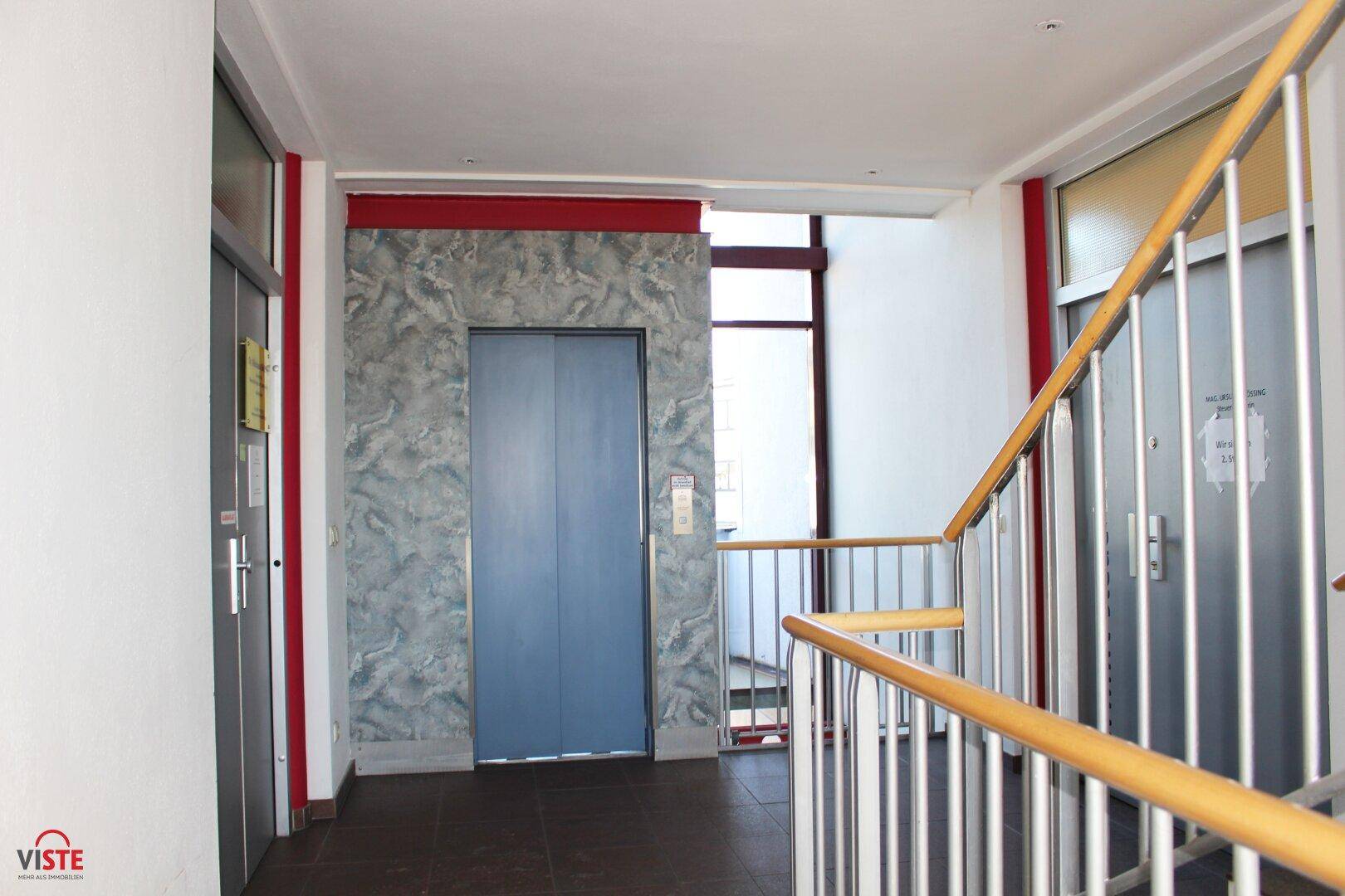 Gemeinschaftsbereich mit Aufzug und Treppenhaus, gekennzeichnet durch eine blaue Aufzugstür und Holzgeländer.