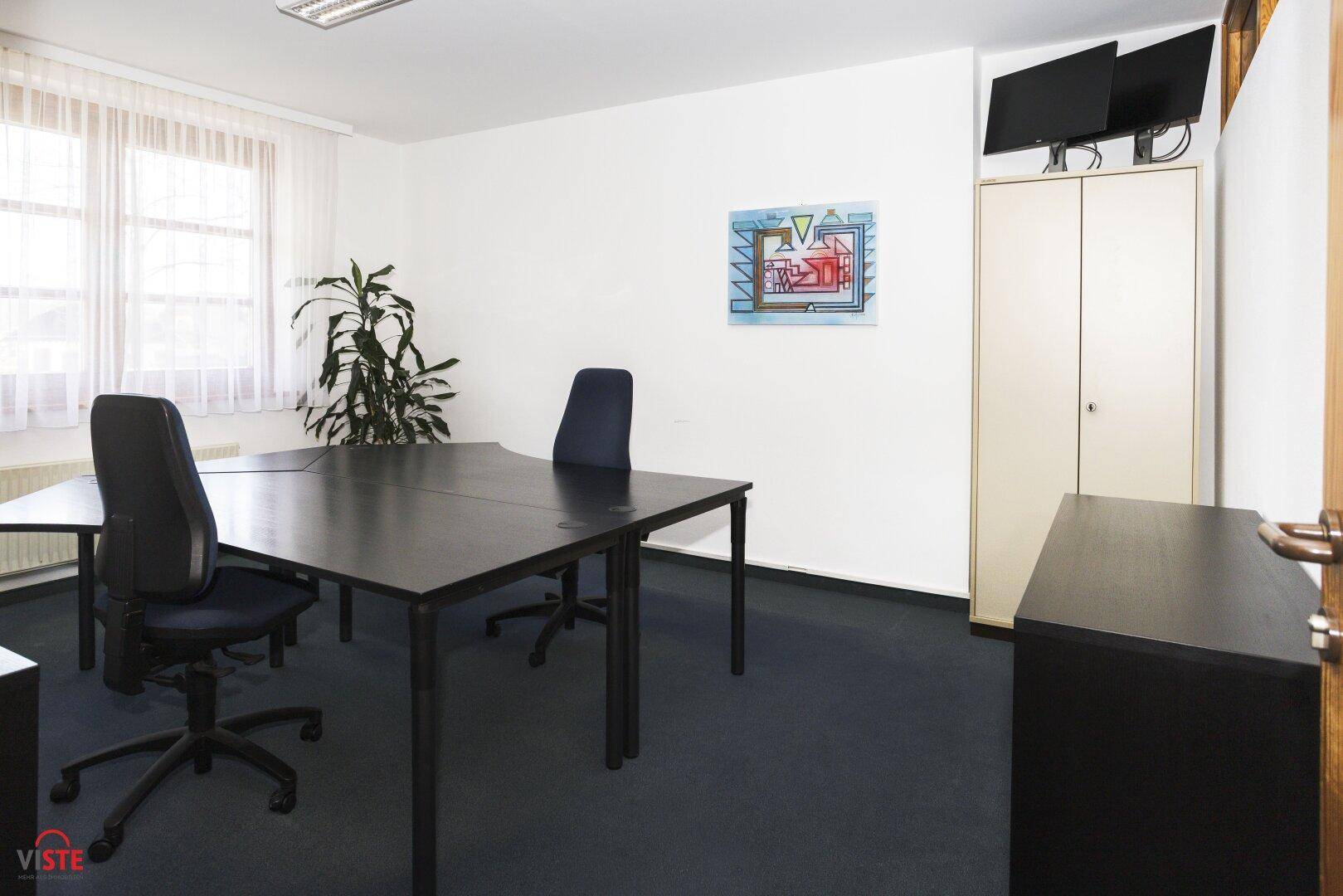Büro mit dunklem Teppichboden, weißen Wänden und einem Schrank mit Monitoren an der Wand.