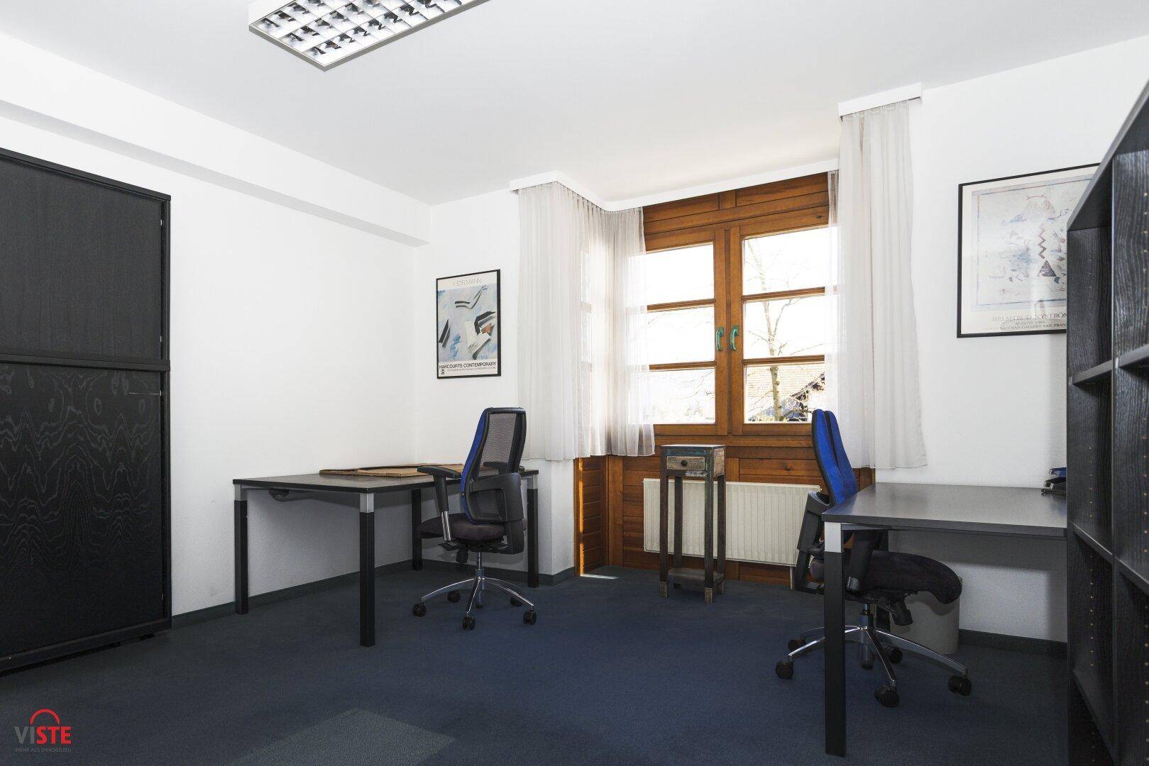 Helles Büro mit zwei Arbeitsplätzen, Holzfensterrahmen und einem dunklen Teppichboden.
