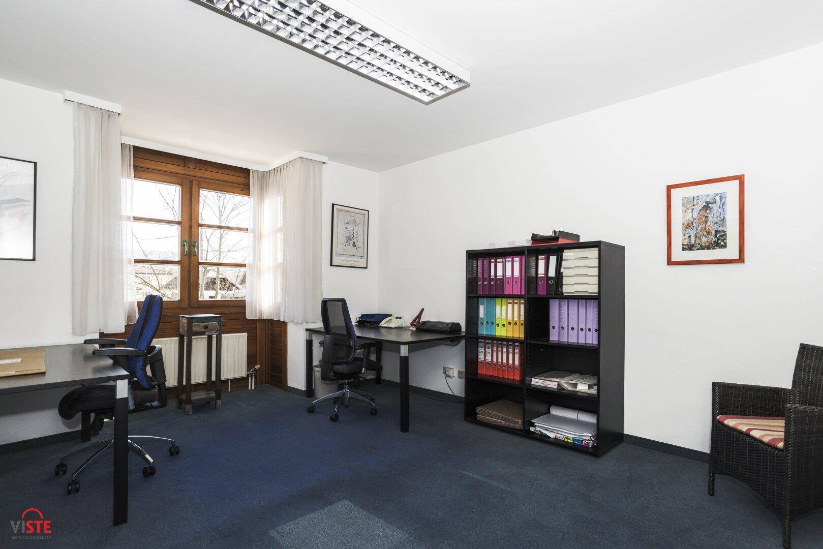 Büro mit mehreren Arbeitsplätzen, einem Bücherregal voller Ordner und einem bequemen Sessel.