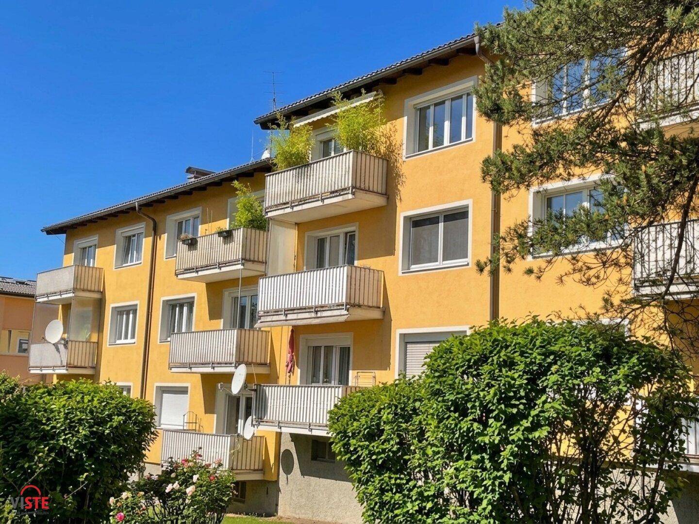 Gelbe Fassade eines Mehrfamilienhauses mit mehreren Balkonen und grüner Bepflanzung.
