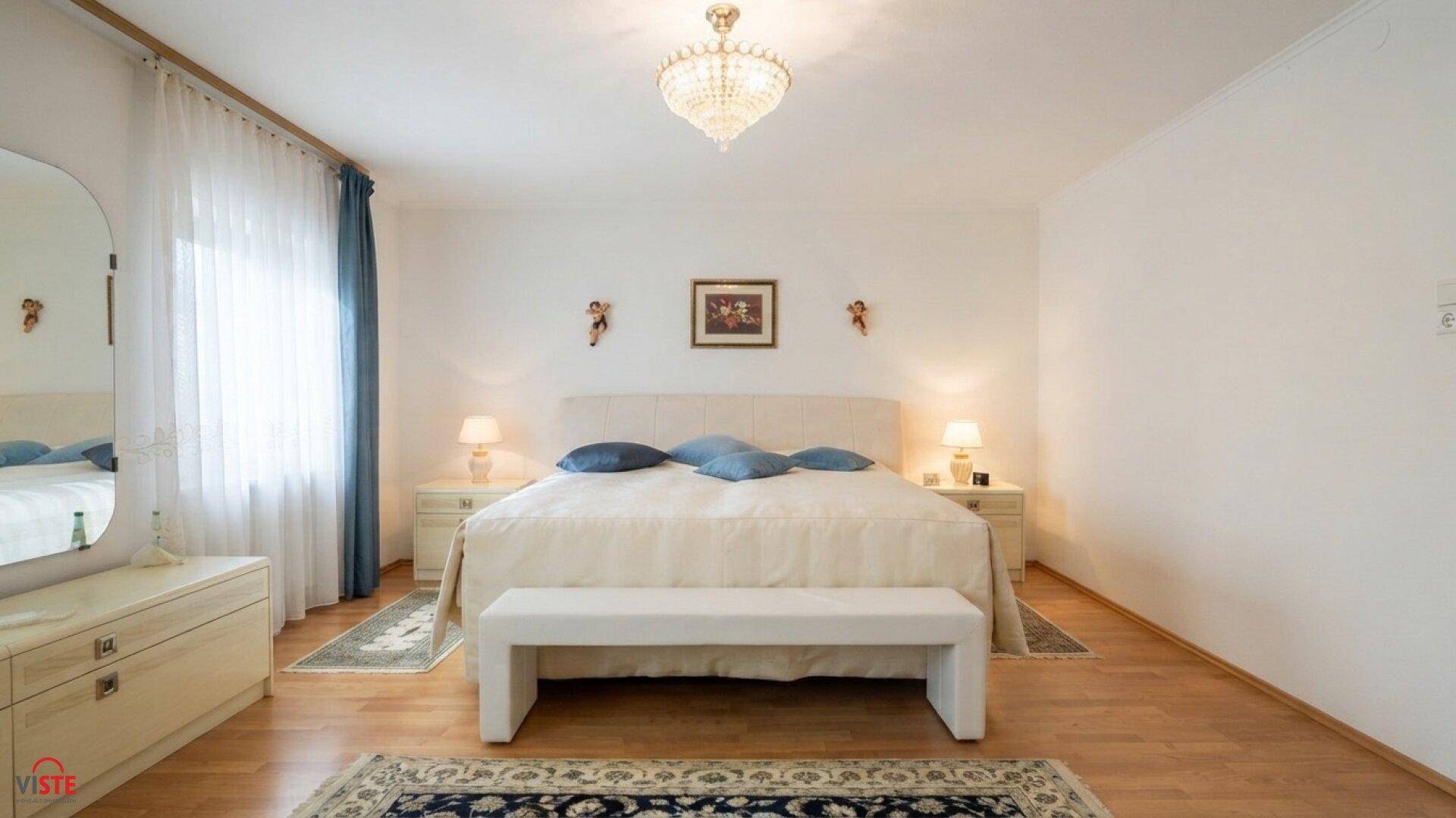 Helles Schlafzimmer mit Doppelbett, Nachttischen und einem Kronleuchter an der Decke.