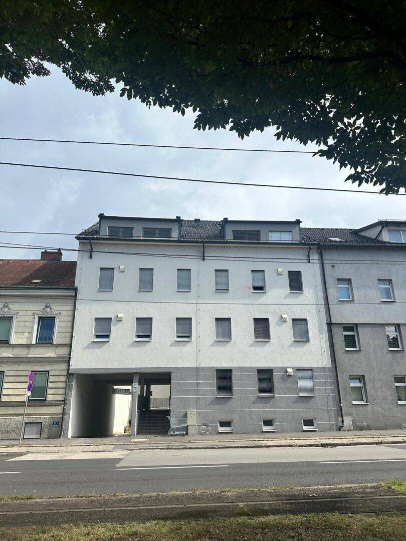 Wohngebäude mit heller Fassade, Rollläden und Dachaufbauten, umgeben von Bäumen.