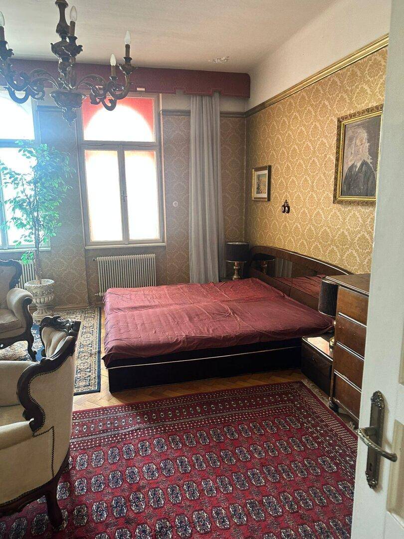 Schlafzimmer mit Doppelbett, klassischem Mobiliar, gemusterter Tapete und Kronleuchter.