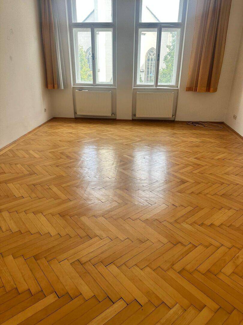 Leeres Zimmer mit Fischgrätparkett, hohen Decken und zwei großen Fenstern.