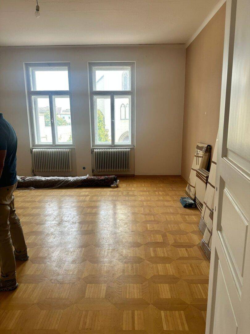 Geräumiges, leeres Zimmer mit Fischgrätparkett und zwei großen Fenstern.