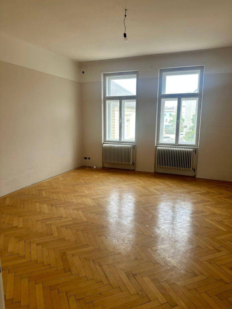 Helles, leeres Zimmer mit Fischgrätparkett und zwei großen Fenstern.