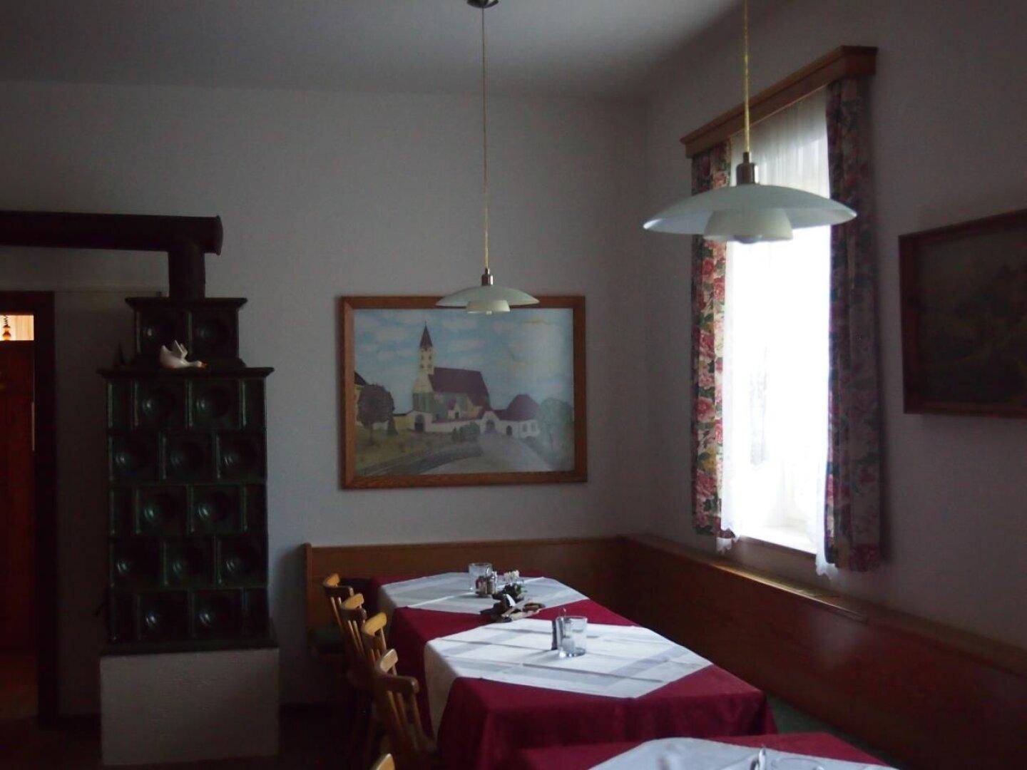 Traditioneller Essbereich mit einem grünen Kachelofen, Holzbänken und einem Fenster mit floralen Vorhängen.