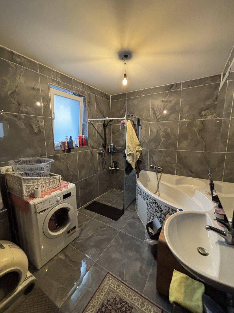 Modernes Badezimmer mit grauen Fliesen, Eckbadewanne, Dusche und Waschmaschine.