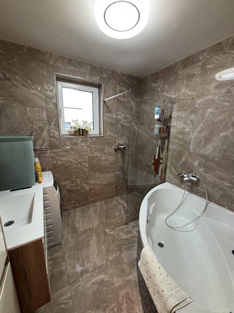 Helles Badezimmer mit braun-grauen Fliesen, Eckbadewanne und Duschbereich.