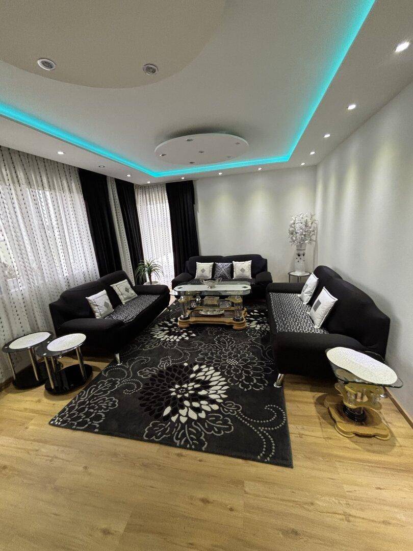 Elegantes Wohnzimmer mit dunklen Sofas, gemustertem Teppich und indirekter Deckenbeleuchtung.