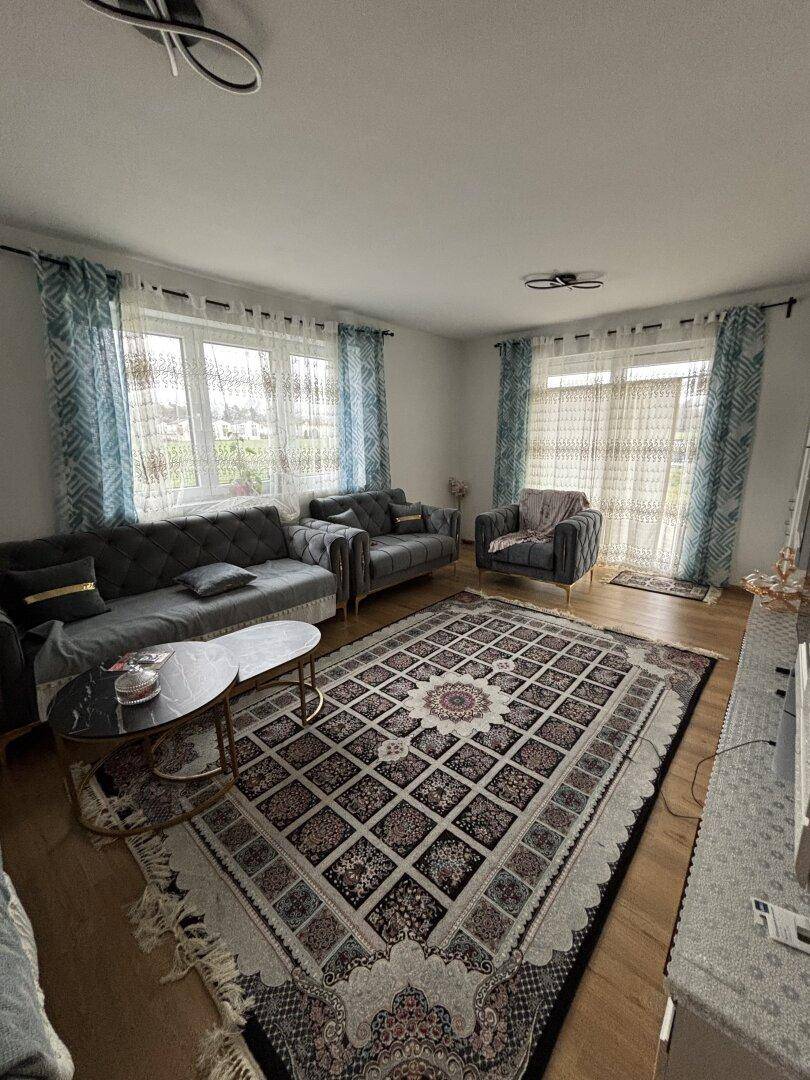 Gemütliches Wohnzimmer mit grauen Sofas, traditionellem Teppich und großen Fenstern.