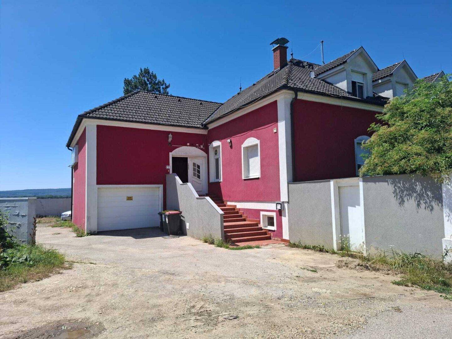 Rotes Gebäude mit Eingangstreppe, Garagentor und gepflastertem Vorplatz.