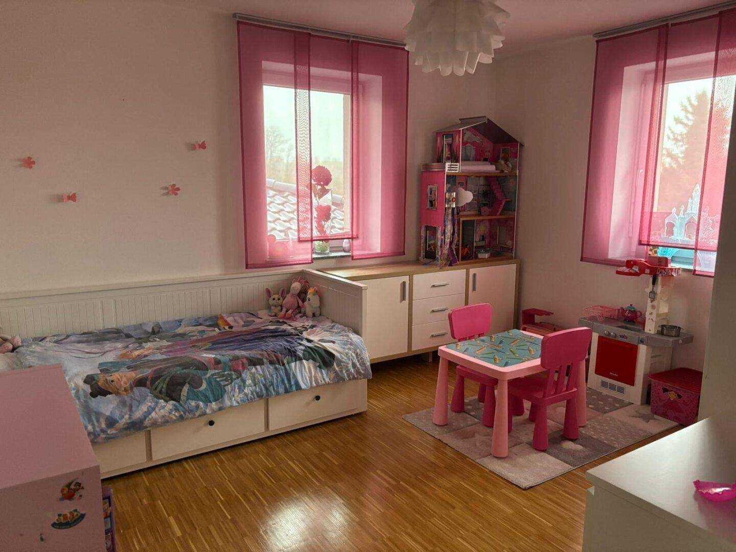 Kinderzimmer mit Holzboden, rosa Vorhängen, Spielzeug und einem Bett mit Stauraum.