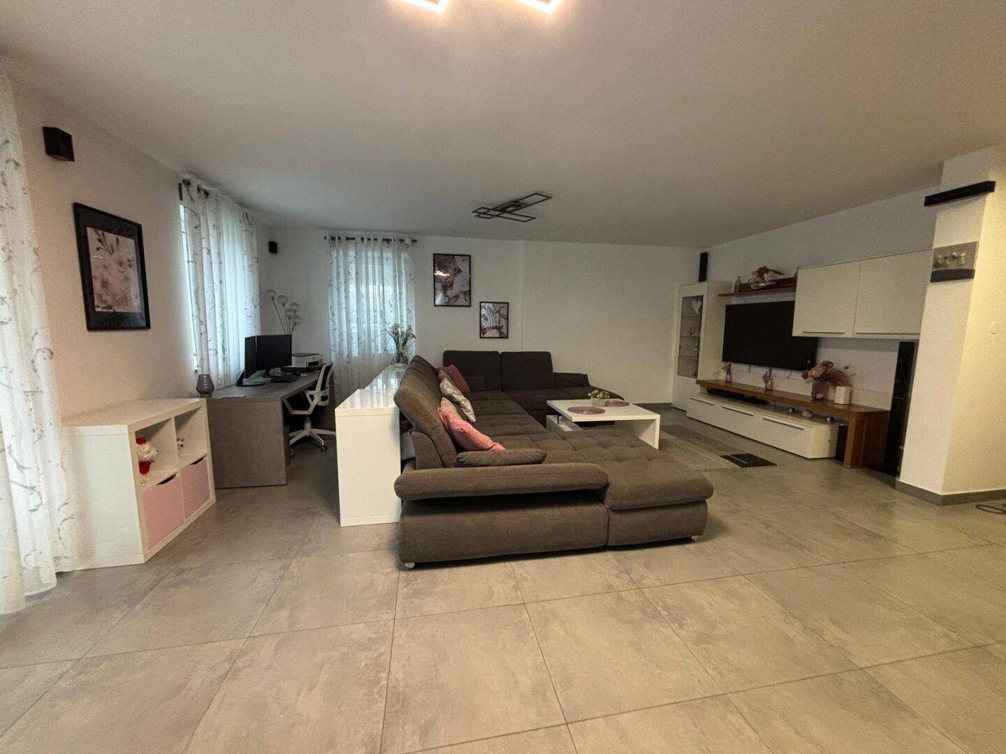 Geräumiges Wohnzimmer mit großem Sofa, Fernseher und Arbeitsbereich im Hintergrund.