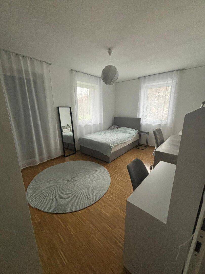 Schlafzimmer mit Einzelbett, Holzfußboden, großem Spiegel und viel Tageslicht.