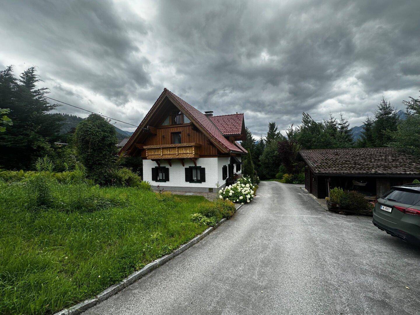 Traditionelles Haus mit Satteldach und Auffahrt, umgeben von Bäumen unter einem dramatischen Himmel.