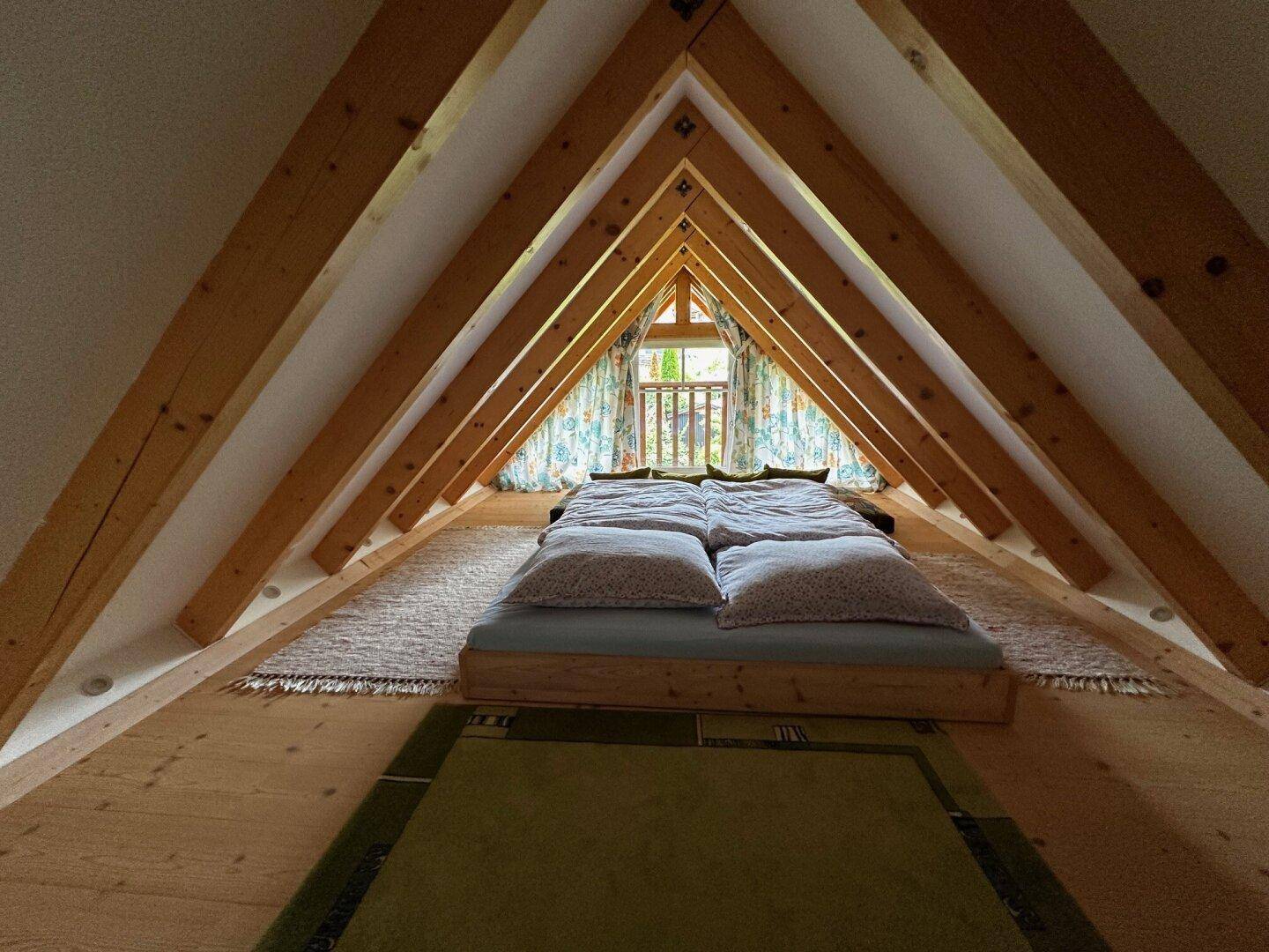 Gemütliches Schlafzimmer im Dachgeschoss mit sichtbaren Holzbalken und Holzboden unter dem Giebelfenster.