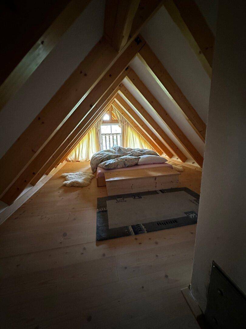 Dachgeschoss-Schlafzimmer mit Holzbalken und einem Bett unter dem Giebelfenster, mit Holzboden.
