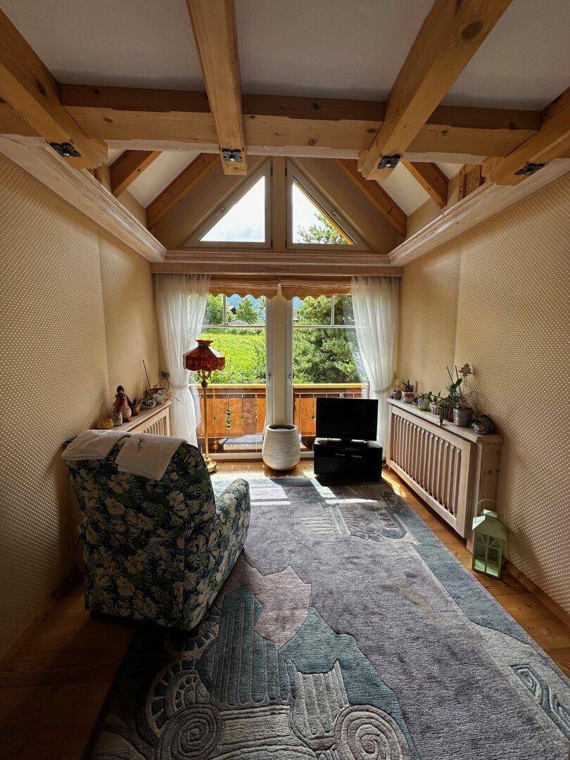Helles Zimmer mit Holzbalkendecke, Teppichboden und großem Fenster mit Blick ins Grüne.