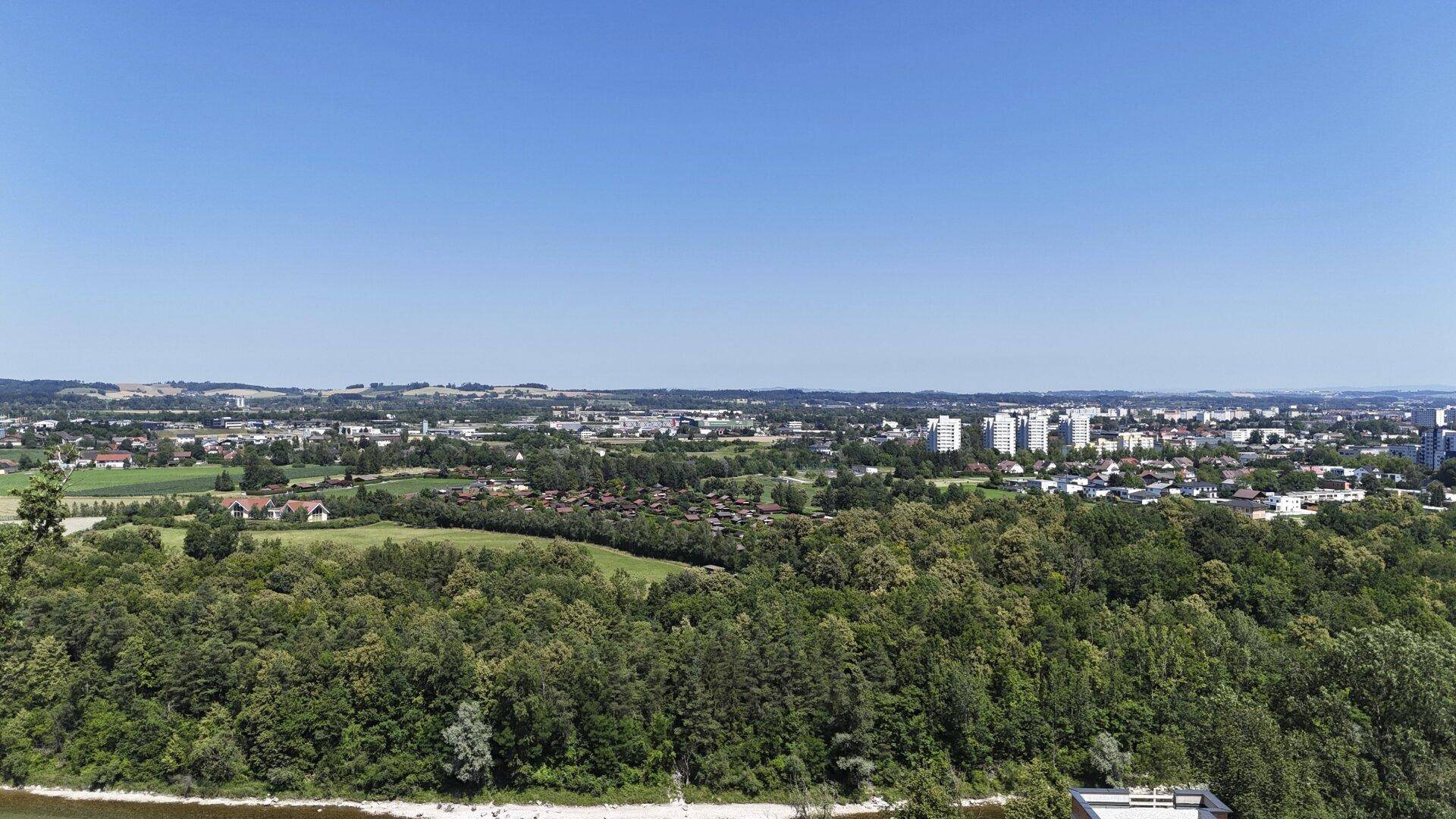 Panoramablick über grüne Wälder, einen Fluss und die Skyline einer entfernten Stadt an einem sonnigen Tag.