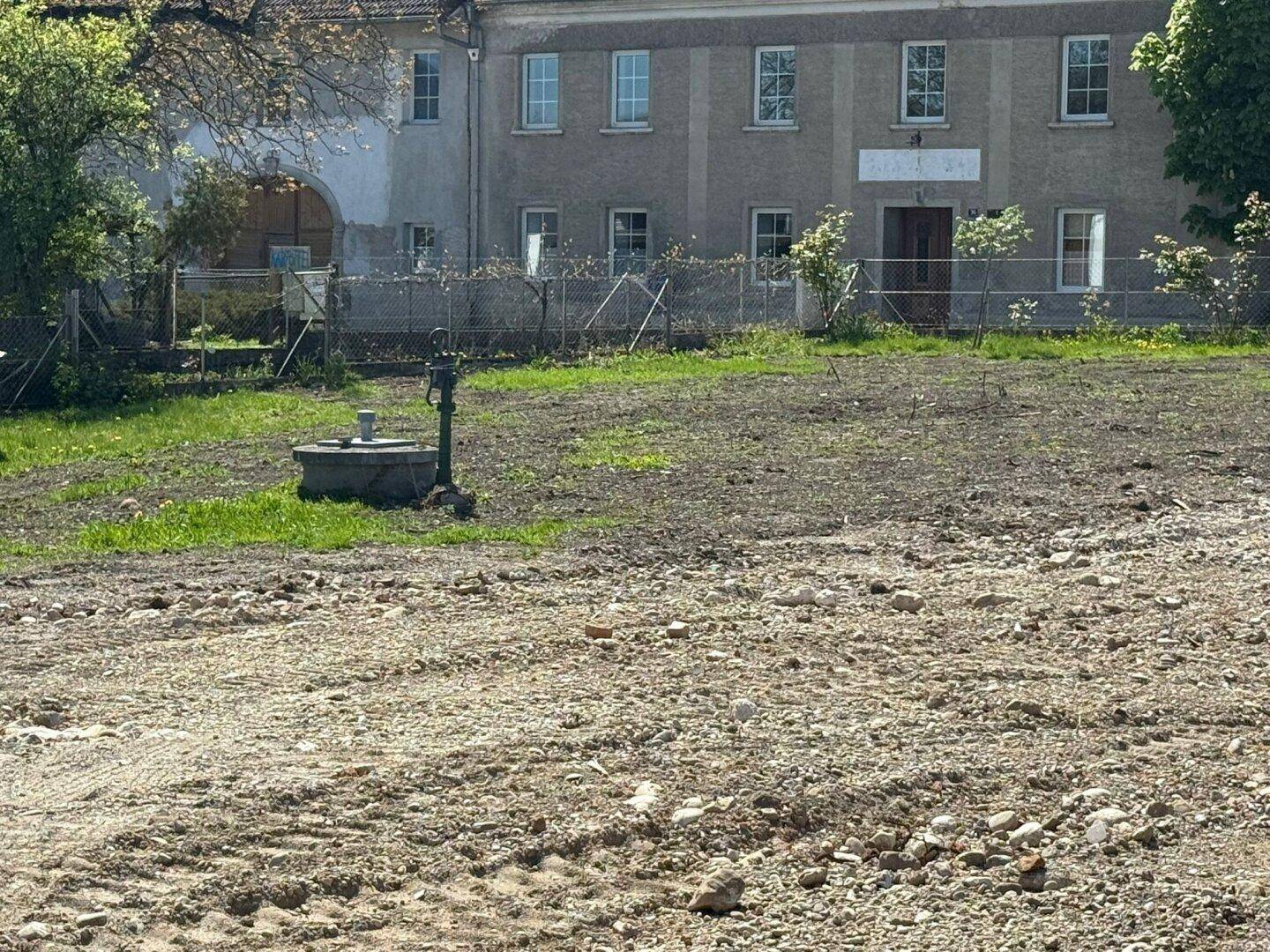 Blick über eine freie Fläche auf ein älteres Gebäude mit mehreren Fenstern und einem alten Brunnen im Vordergrund.