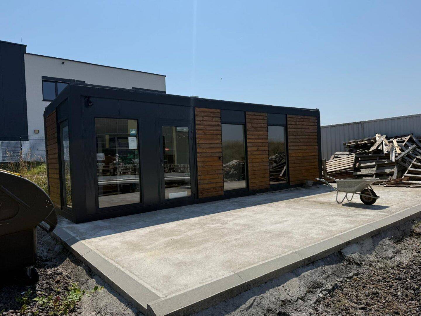 Modernes Modulgebäude mit dunkler Fassade und Holzverkleidung, davor eine gepflasterte Terrasse.