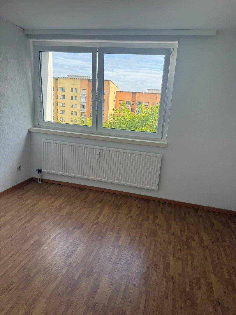 Geräumiges Zimmer mit Parkettboden, großem Fenster und Ausblick auf umliegende Gebäude und Bäume.