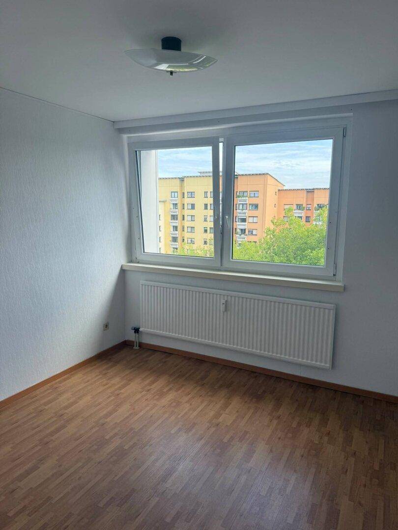 Freundliches Zimmer mit Parkettboden, großem Fenster und Ausblick auf die Wohngebäude.