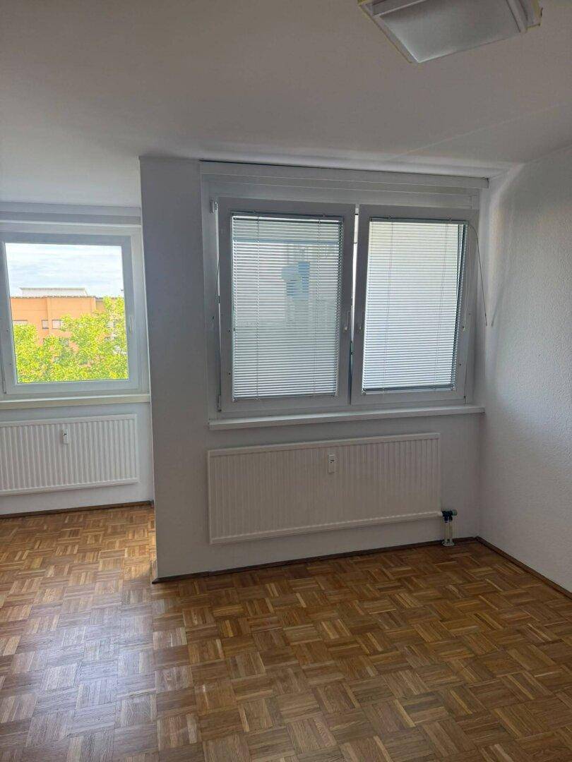 Helles Zimmer mit Parkettboden, Jalousien an den Fenstern und Blick auf die Umgebung.