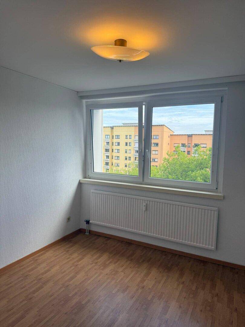 Helles Zimmer mit Parkettboden, Fenster und Blick auf die umliegenden Gebäude und Bäume.