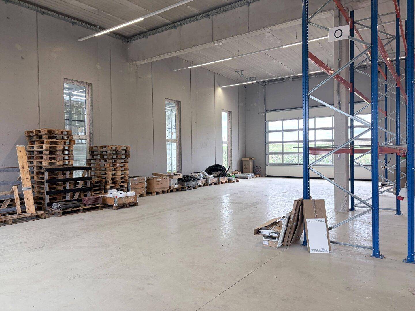Leere Lagerhalle mit hohen Betonwänden, hohen Fenstern und einem großen Rolltor, mit gestapelten Paletten.