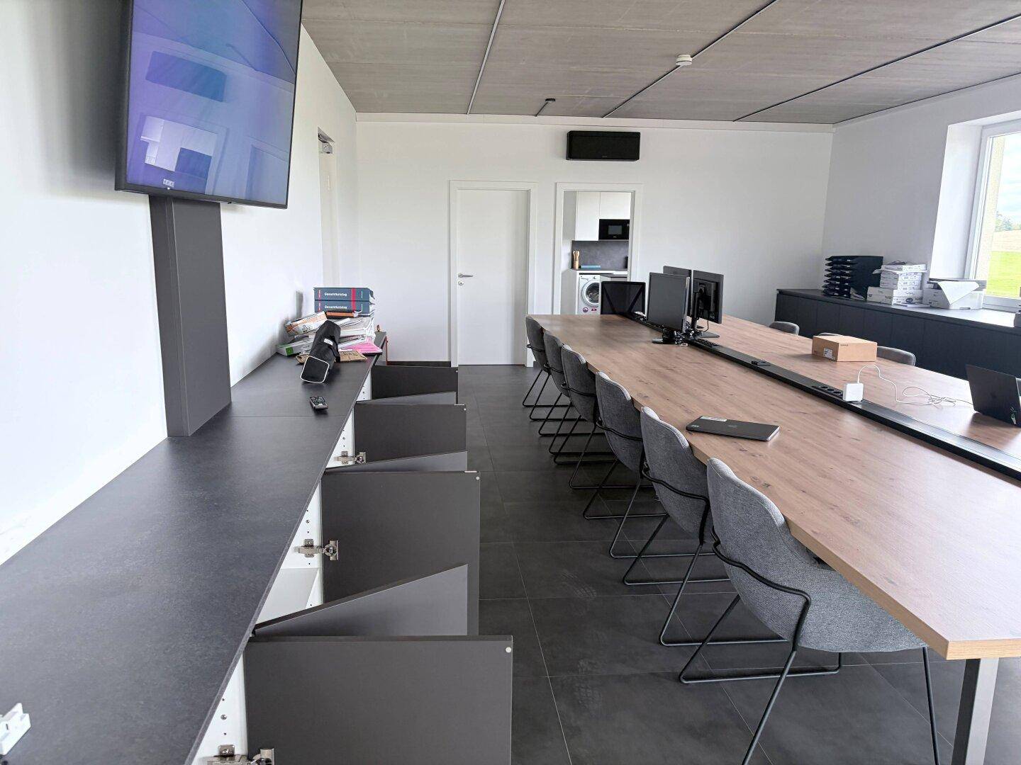 Moderner Konferenzraum mit langem Tisch, Stühlen, Monitoren und einem Wandfernseher.