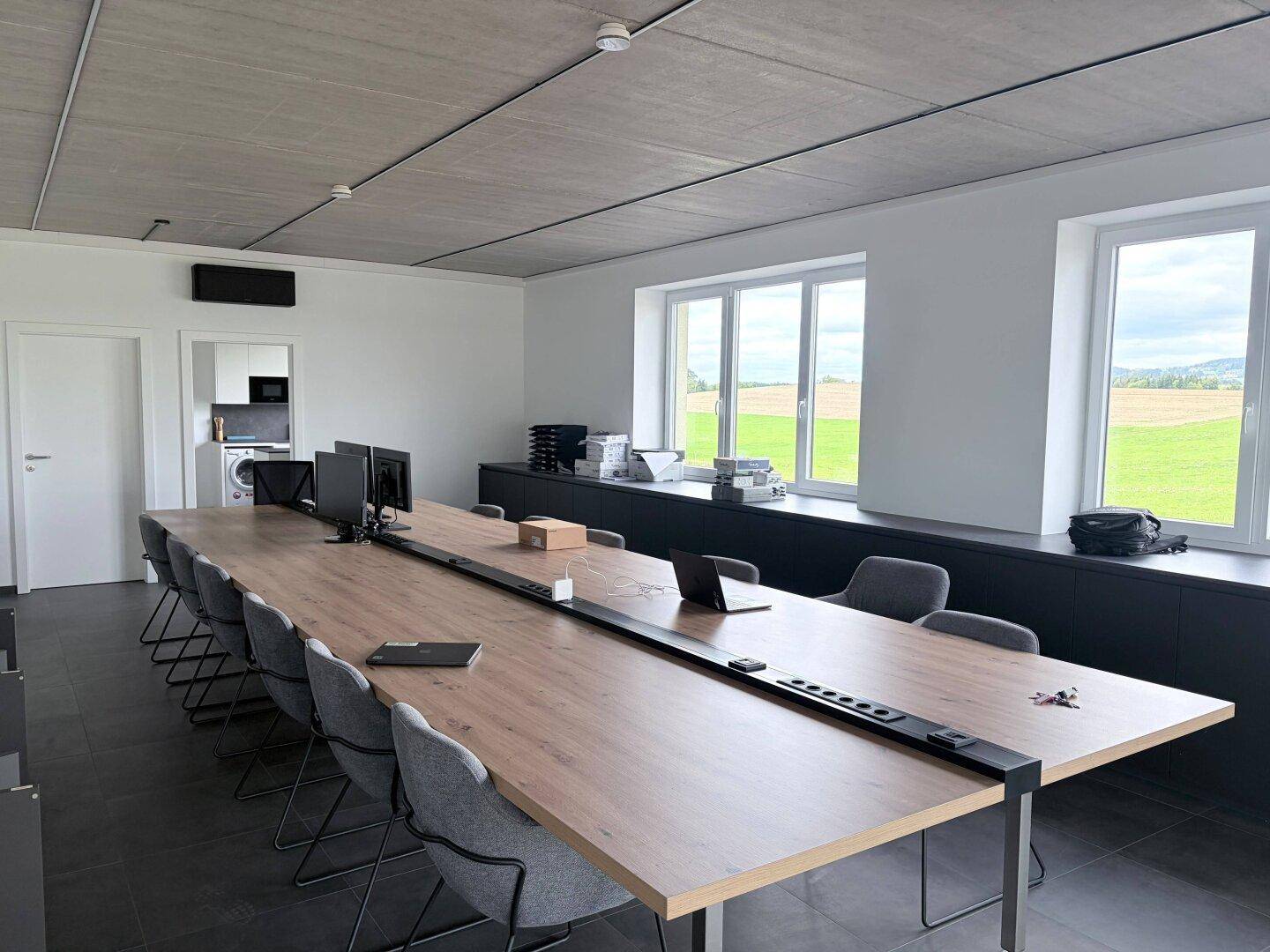 Weitwinkelaufnahme des modernen Konferenzraums mit langem Tisch, Stühlen und Fenstern mit grüner Aussicht.