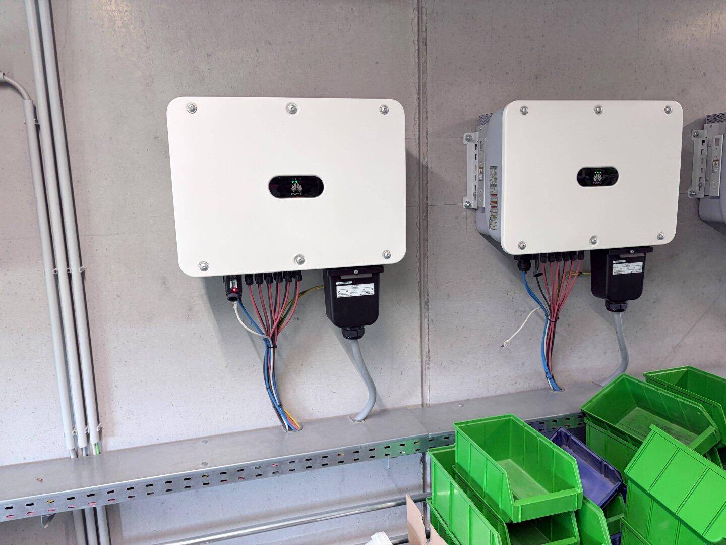 Zwei Wechselrichter für eine Photovoltaikanlage sind an der Wand montiert, mit grünen Behältern darunter.