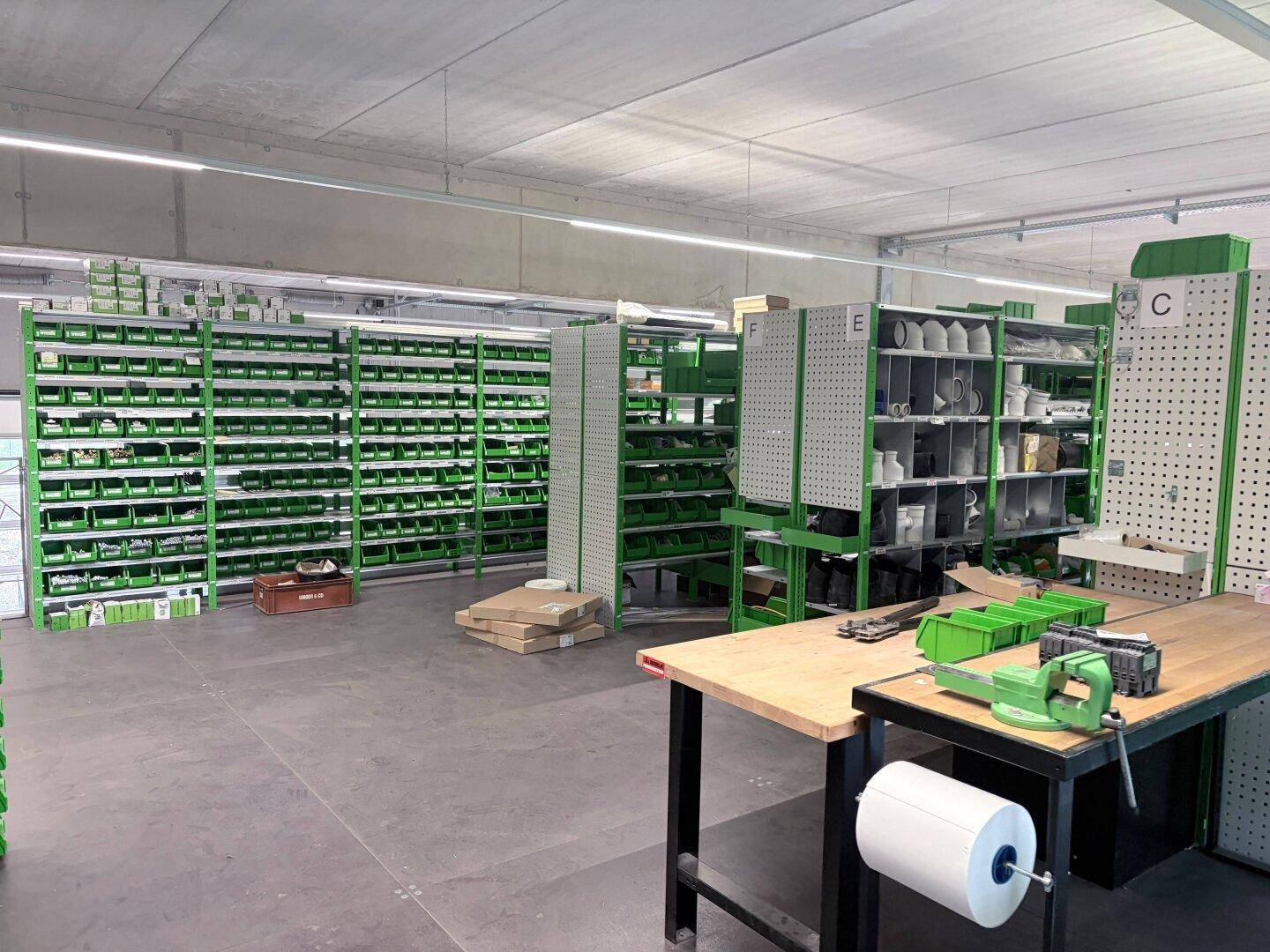Lagerraum mit grünen Regalen voller kleiner Behälter und einem Arbeitstisch mit Schraubstock.