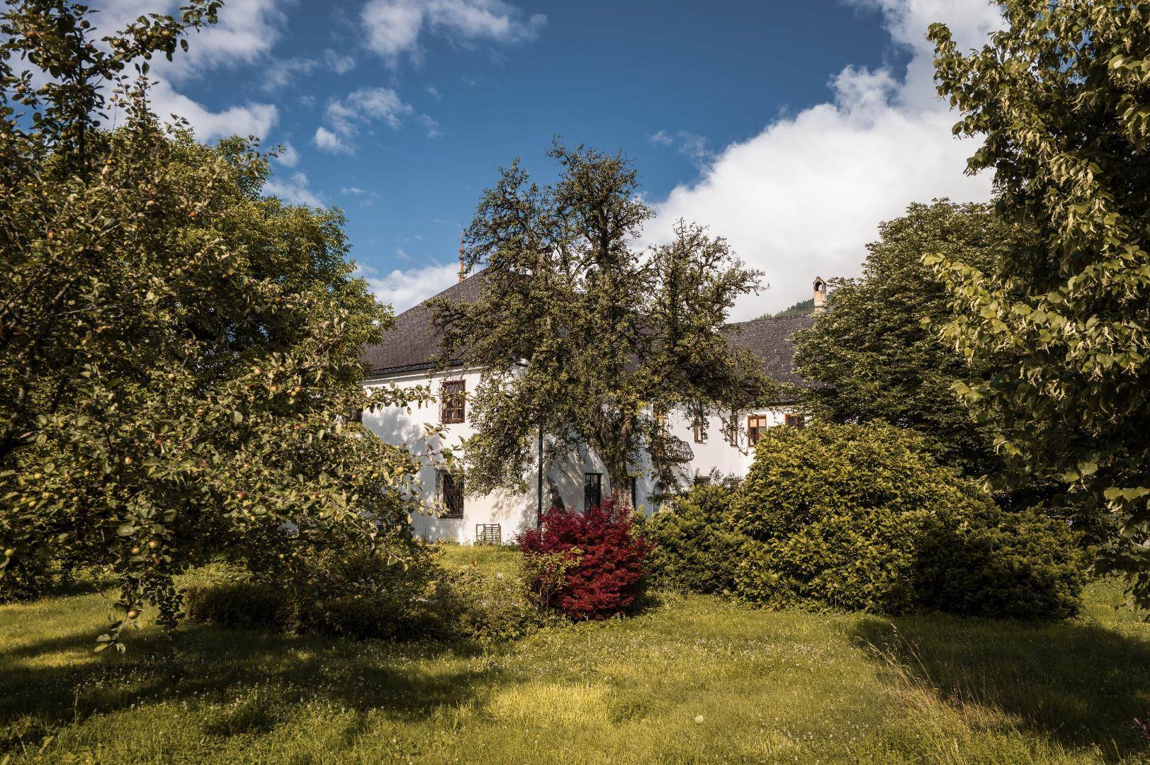 Weitläufiger Garten mit altem Baumbestand und einem historischen Gebäude im Hintergrund unter blauem Himmel.