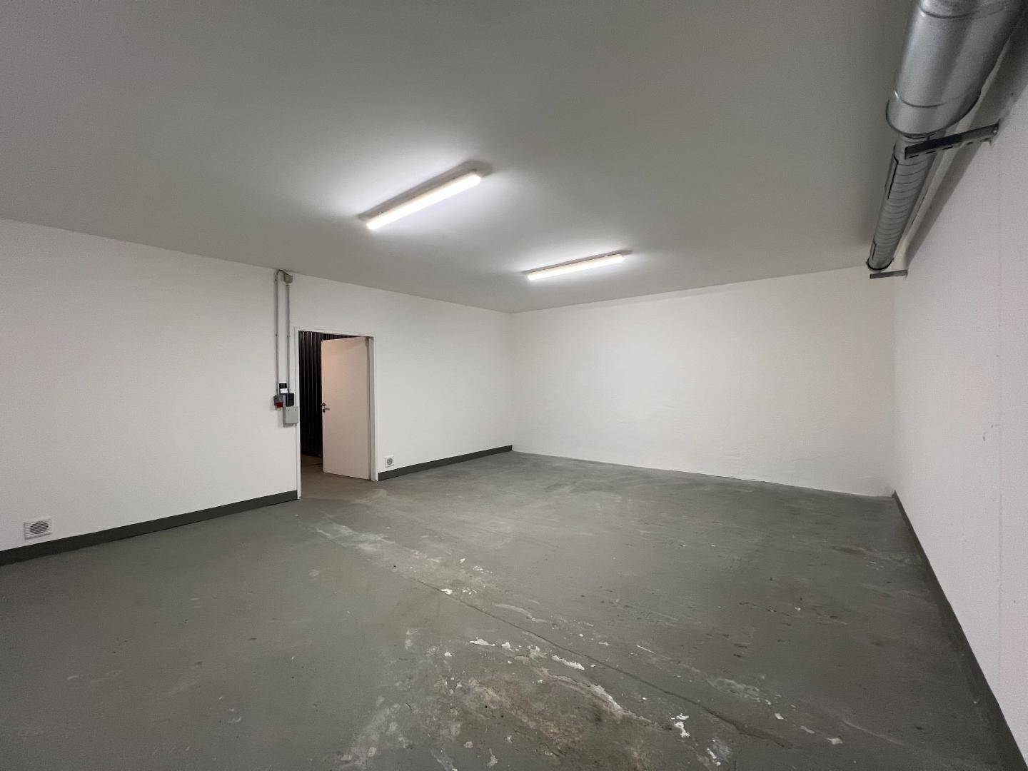 Heller Lagerraum mit weiß gestrichenen Wänden, Betonboden und offenliegenden Deckenrohren.